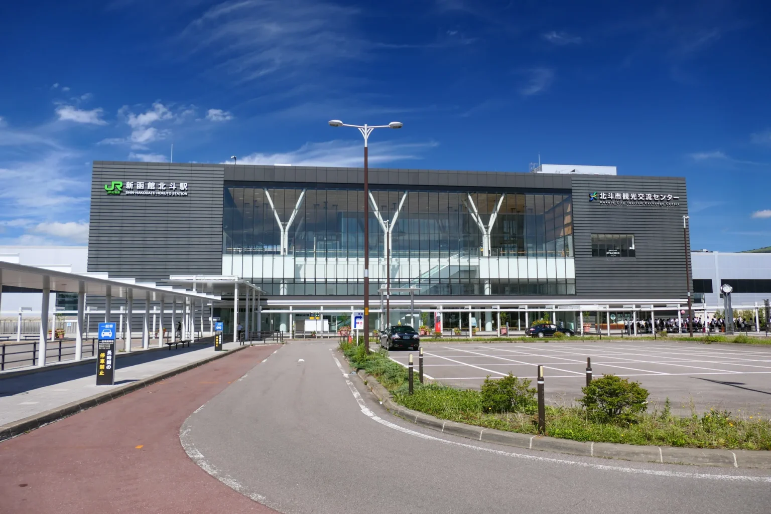 The glass-fronted south exit building of Shin-Hakodate-Hokuto Station, with the JR station name sign on the left block and the Hokuto City Tourism Exchange Center sign on the right, under a blue sky.