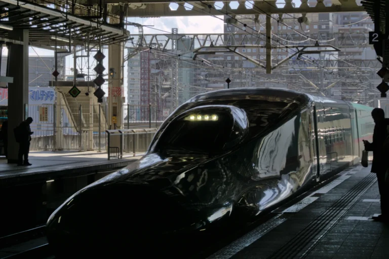 An E5 series shinkansen, its long glossy nose in shadow and headlights lit, stands at a platform of Sendai Station under the steel canopy, with overhead wires and city buildings behind.