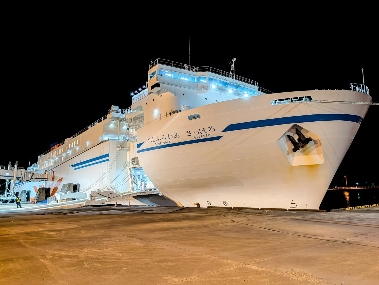 The white bow of the ferry Sunflower Sapporo, its name painted in Japanese and English with a blue stripe, moored at night beside the Oarai ferry terminal with lights on along its decks.