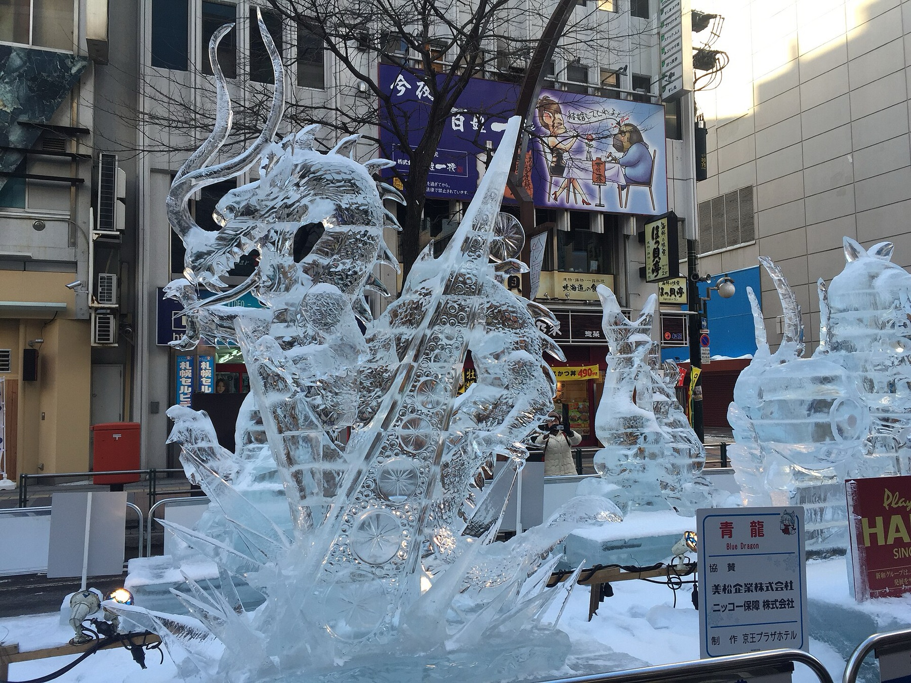 A crystal-clear ice sculpture of a dragon named Blue Dragon standing on the Ekimae-dori street at the Susukino site of the Sapporo Snow Festival, with its official name plate in Japanese and English at its base.