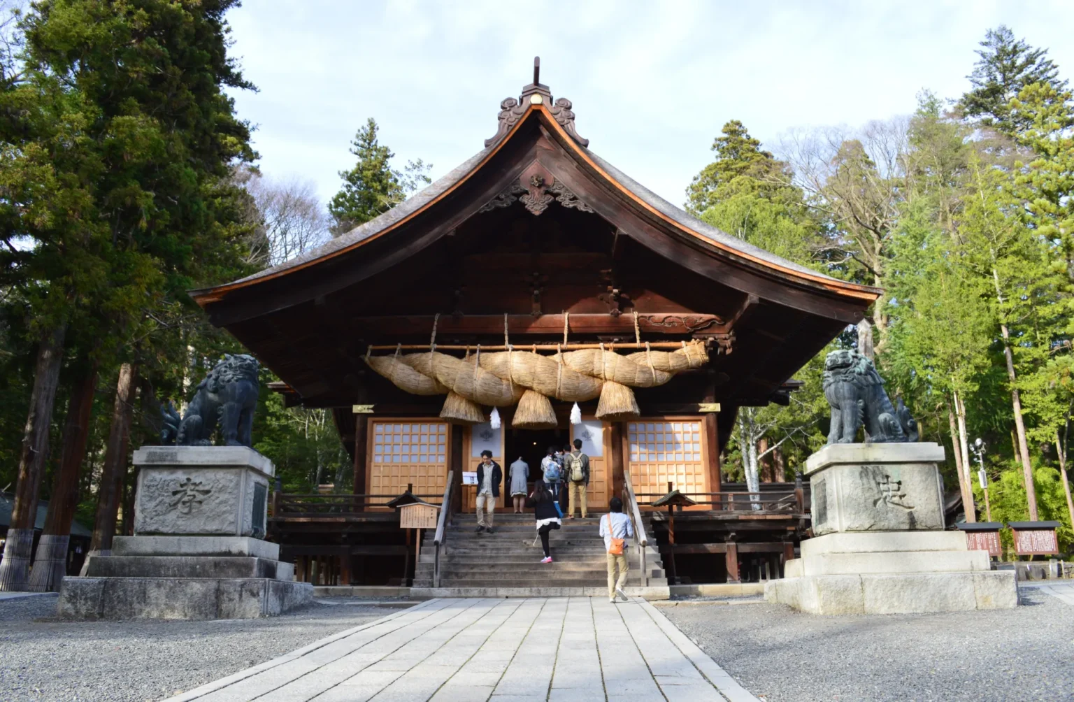 The kagura hall at Suwa Taisha Shimosha Akimiya: a dark timber hall with a deeply curved roof, hung with an enormous braided rice-straw shimenawa rope, flanked by two bronze guardian lions on stone plinths, with visitors climbing the stone steps.