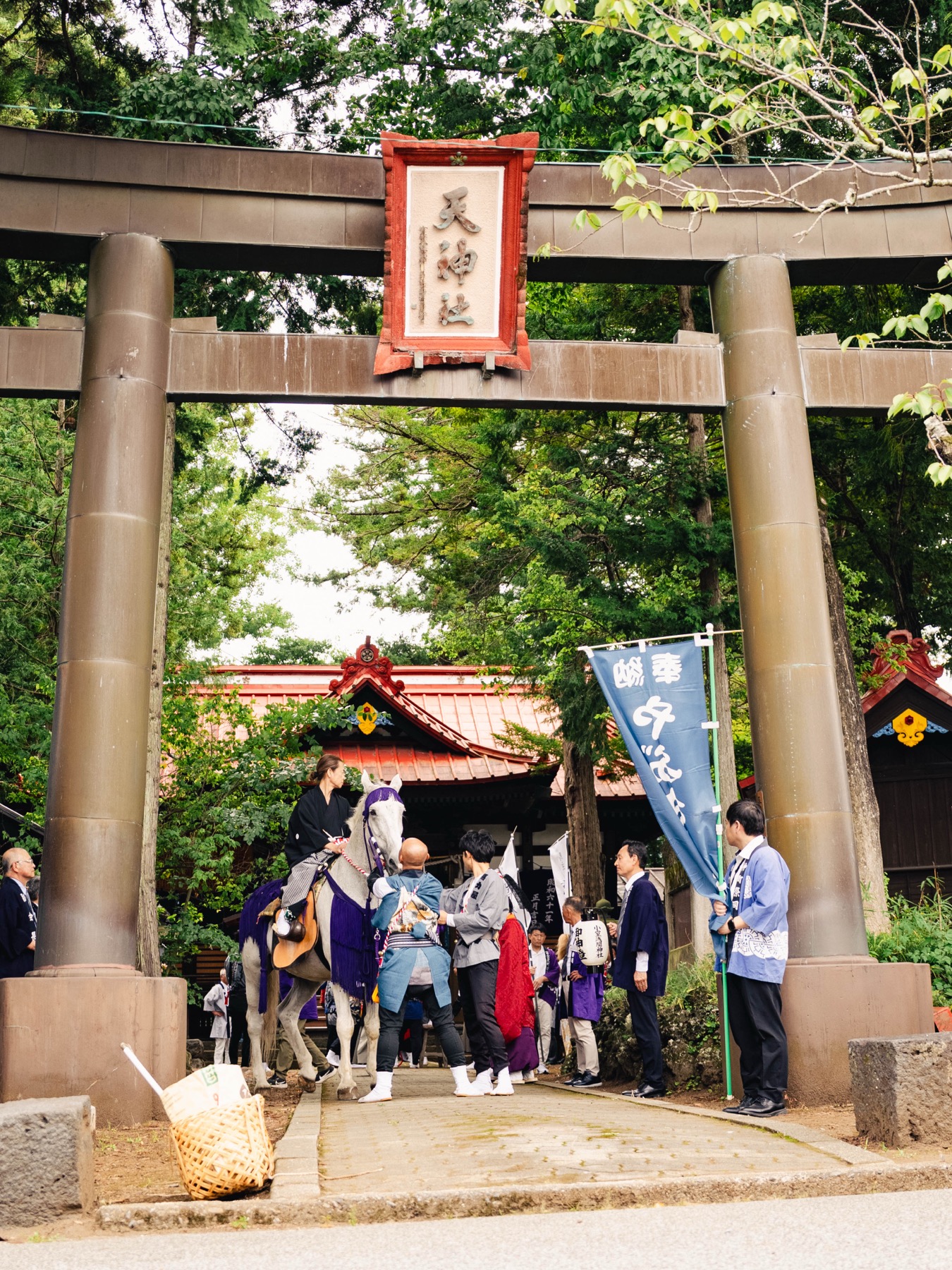 The yabusame procession pauses inside the stone torii gate of Tenjinsha in Fujiyoshida, the white horse and attendants visible under the gate plaque and a blue festival banner