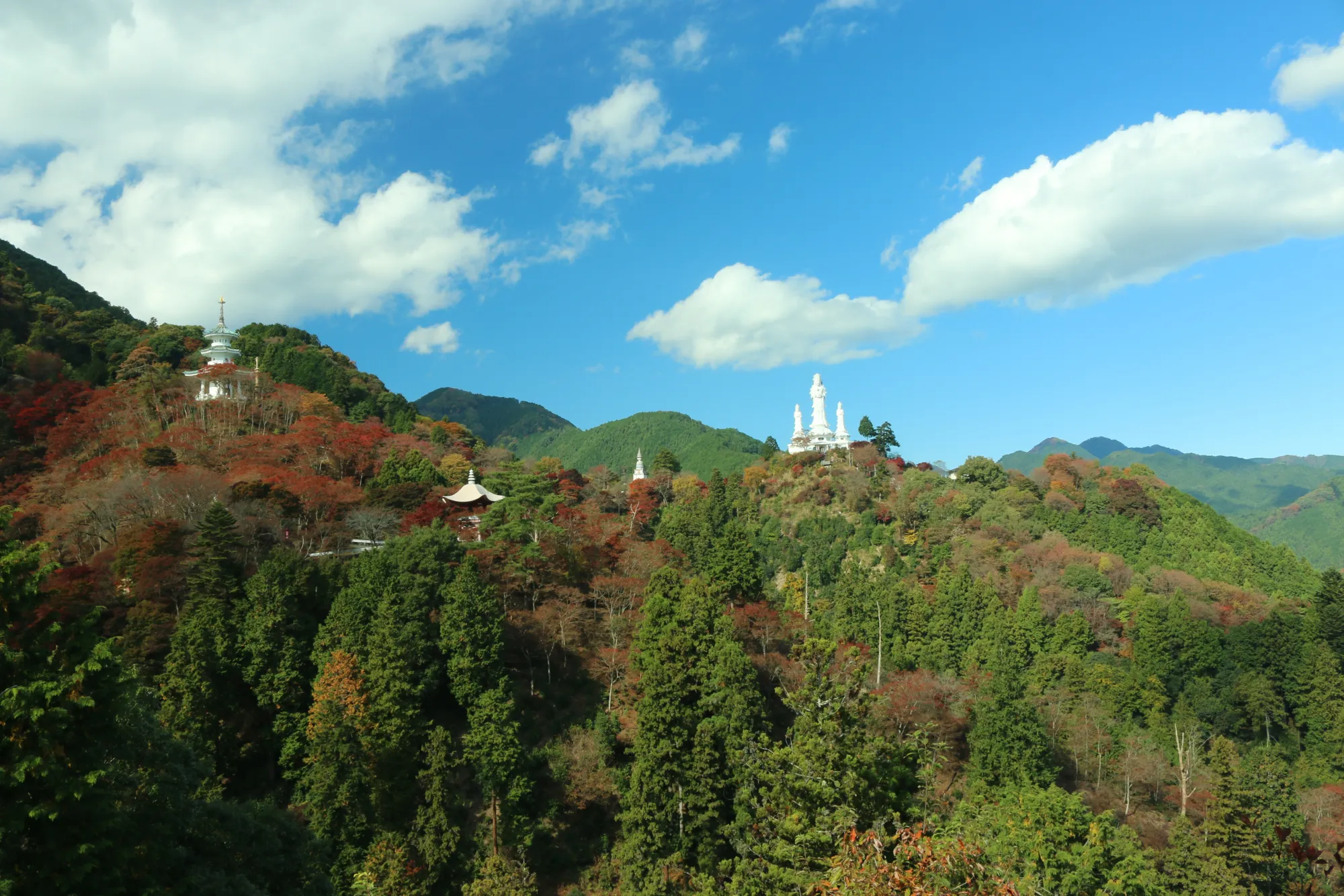 A wide view of Hakuunzan Torii Kannon in Hanno, Saitama: a white Kannon statue standing on the ridge at right, a white pagoda-shaped tower on a spur at left, and further roofs and structures scattered down a wooded mountainside patched with early autumn colour under a blue sky.
