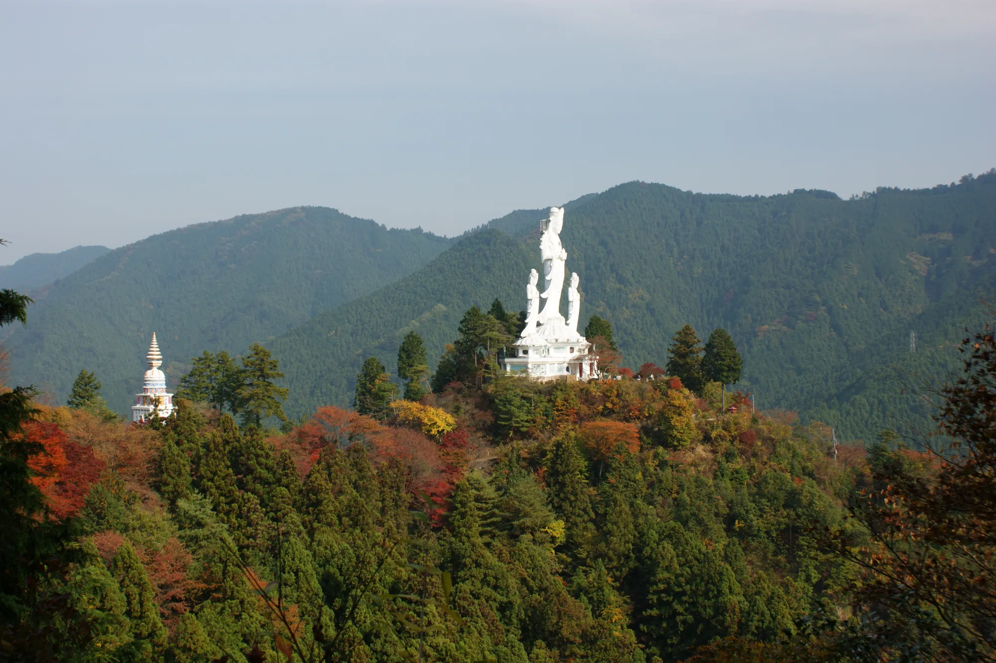 The white Kusei Dai-Kannon at Torii Kannon standing on a stone base on a forested ridge, flanked by two attendant figures of similar height, with the white Xuanzang Sanzo tower visible on a separate spur to the left and autumn colour along the slope below.