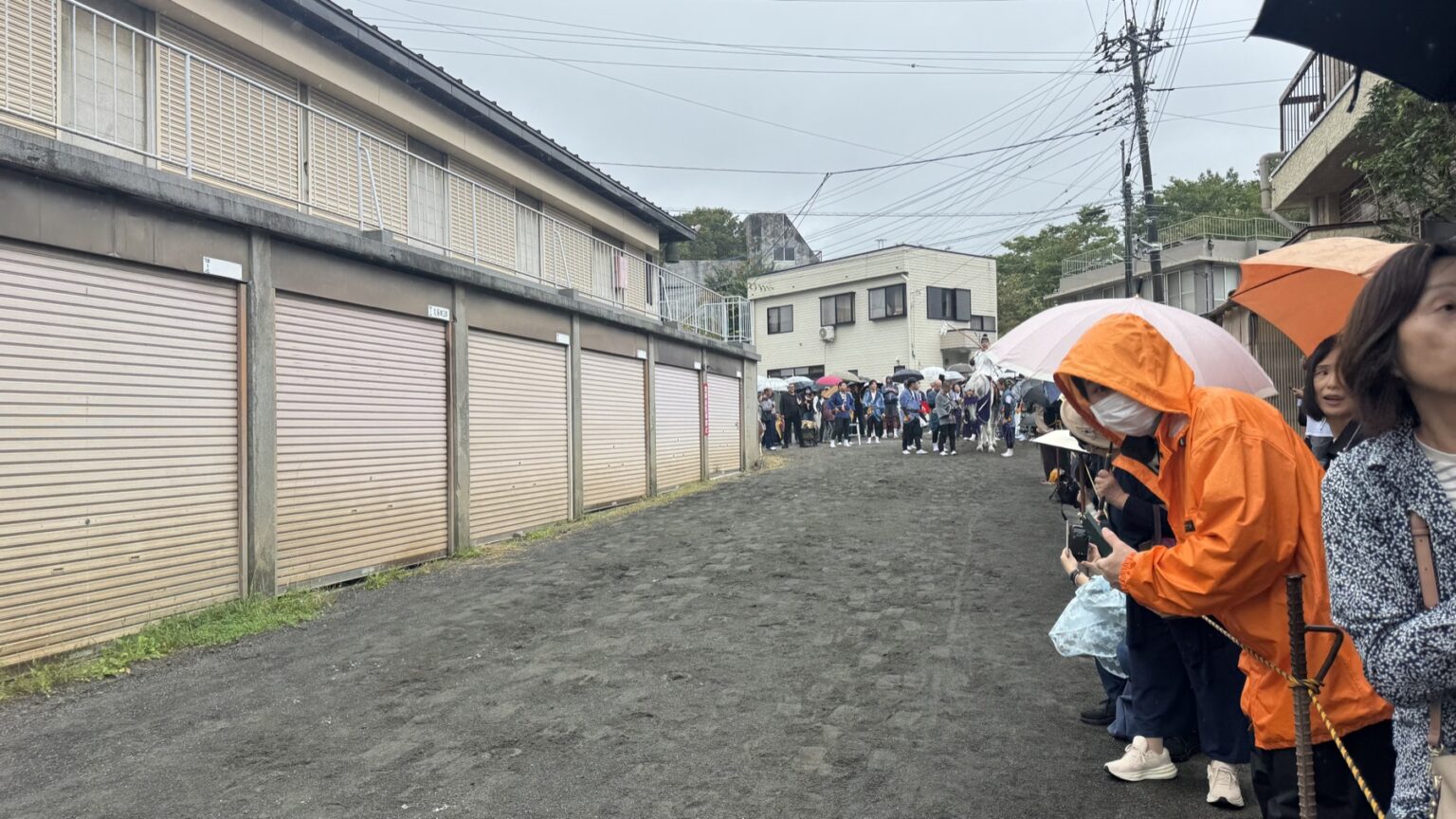 Spectators in raincoats and under umbrellas line the packed-sand lane between lock-up garages and houses, waiting for the yabusame runs at Omuro Sengen Shrine in Shimoyoshida
