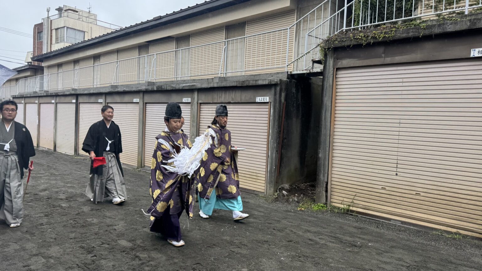 Two men in purple robes patterned with gold chrysanthemums carry white paper wands past formally dressed shrine officials in black haori and hakama at the Fujiyoshida yabusame festival
