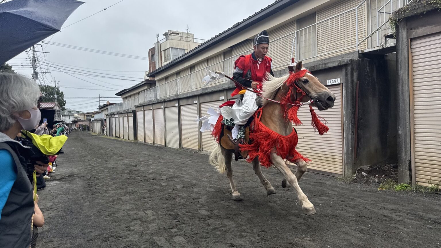 A rider in scarlet ritual dress gallops a chestnut horse hung with red tassels down the narrow garage-lined course at the Fujiyoshida yabusame festival in the rain, with spectators under umbrellas at the roadside