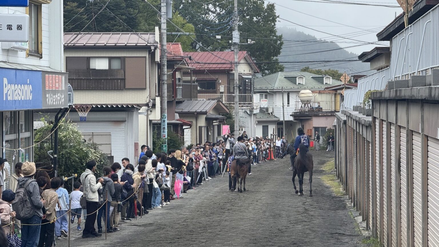 Two festival horses and their riders move down Honcho-dori, the retro shopping street in Fujiyoshida lined with an old Panasonic sign and shuttered shopfronts, with spectators behind ropes on a grey rainy day