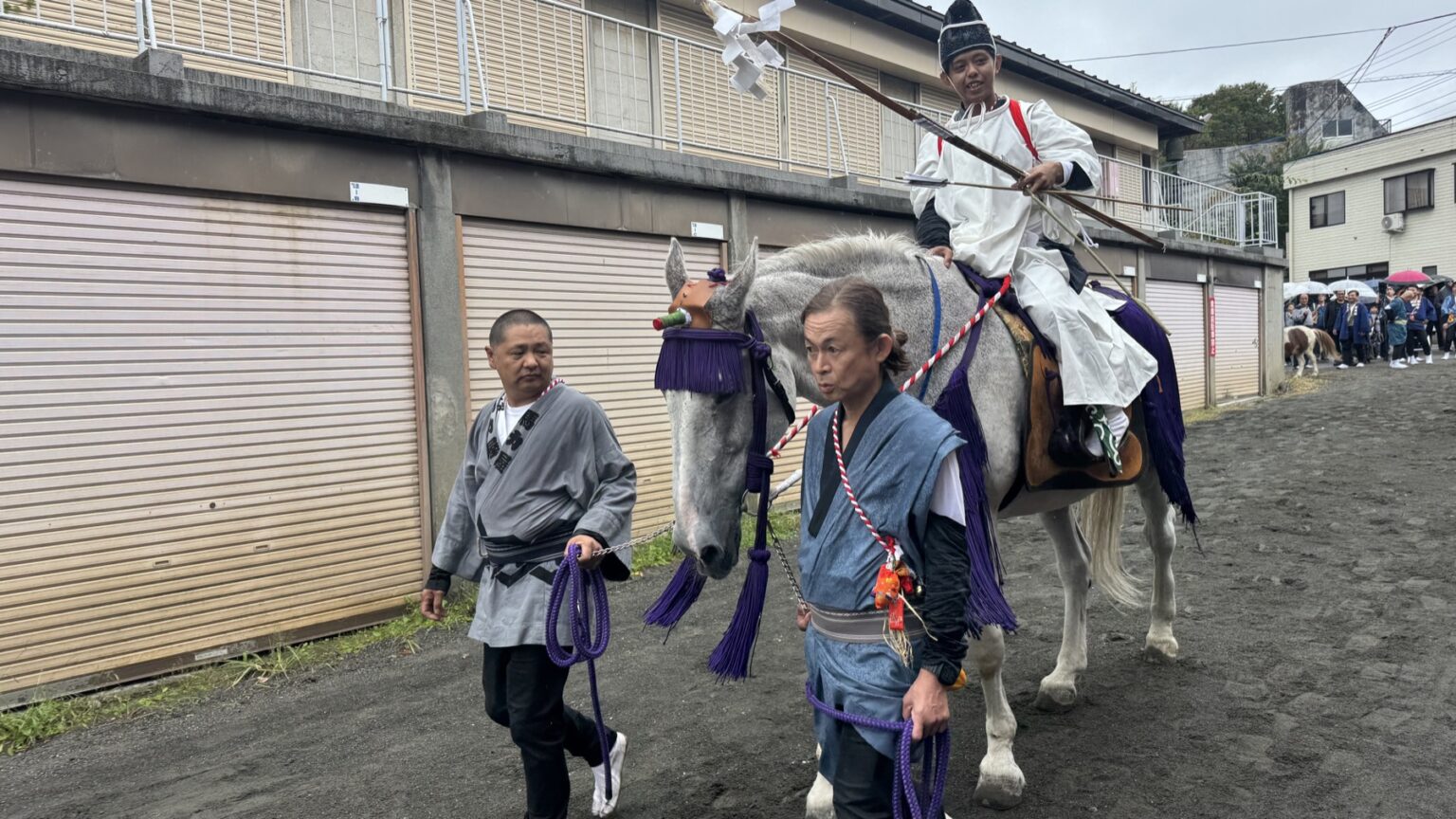 A white shrine horse in purple tassels, led by two handlers in indigo happi coats, carries a rider in white ritual dress holding a bow along the yabusame course at Omuro Sengen Shrine in Shimoyoshida