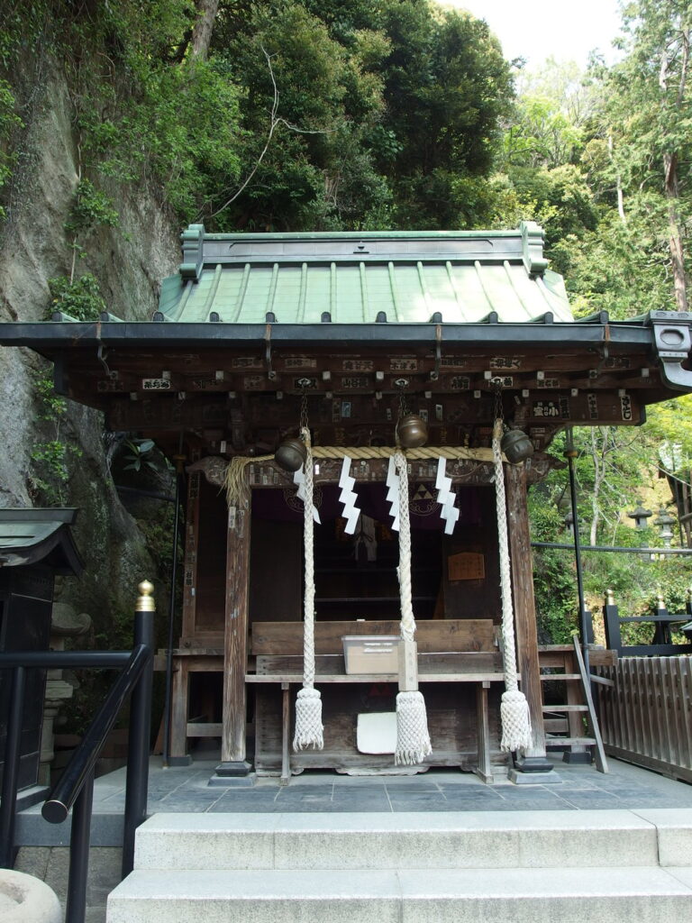 The main hall of Zeniarai Benten Ugafuku Shrine standing against the cliff wall inside its bowl of rock, with a straw shimenawa rope across the cave shrine