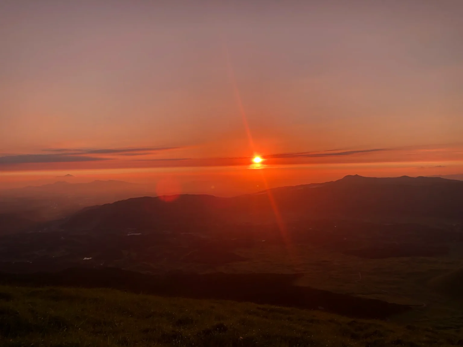 An orange sunset with a bright sun and lens flare above a dark line of hills; below, the darkening caldera floor with scattered buildings and fields.