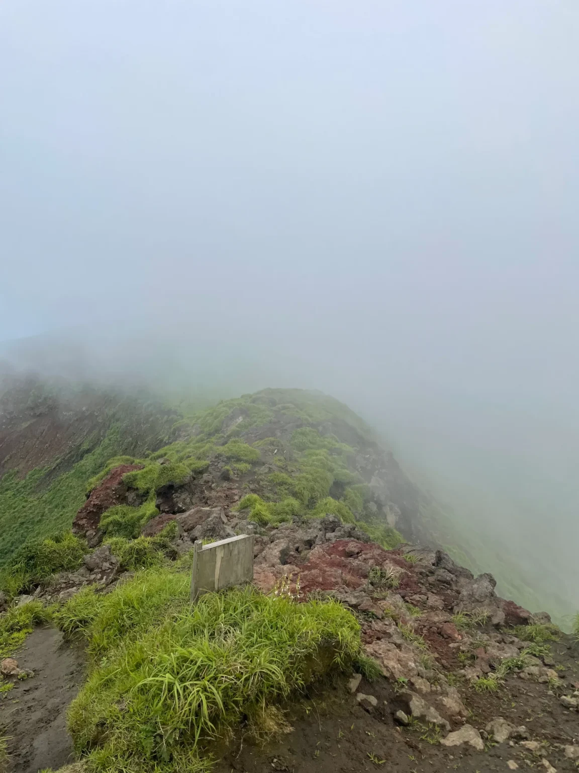 Looking down a narrow ridge of red and dark volcanic rock and tufts of grass, with a small grey wooden signboard beside the path and fog hiding everything beyond.