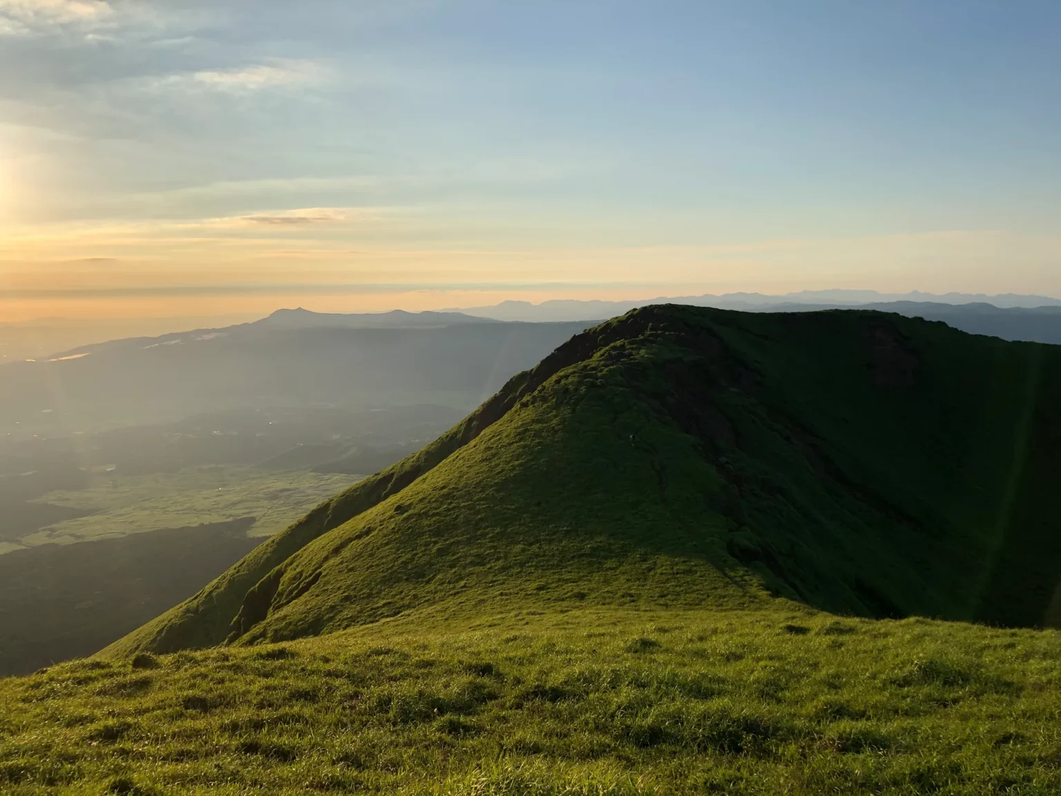 A grass-covered ridge rising to a peak in low evening sun, its right flank dropping steeply with dark rock showing; on the left, the caldera floor in haze and a distant flat-topped mountain under a pale sky.