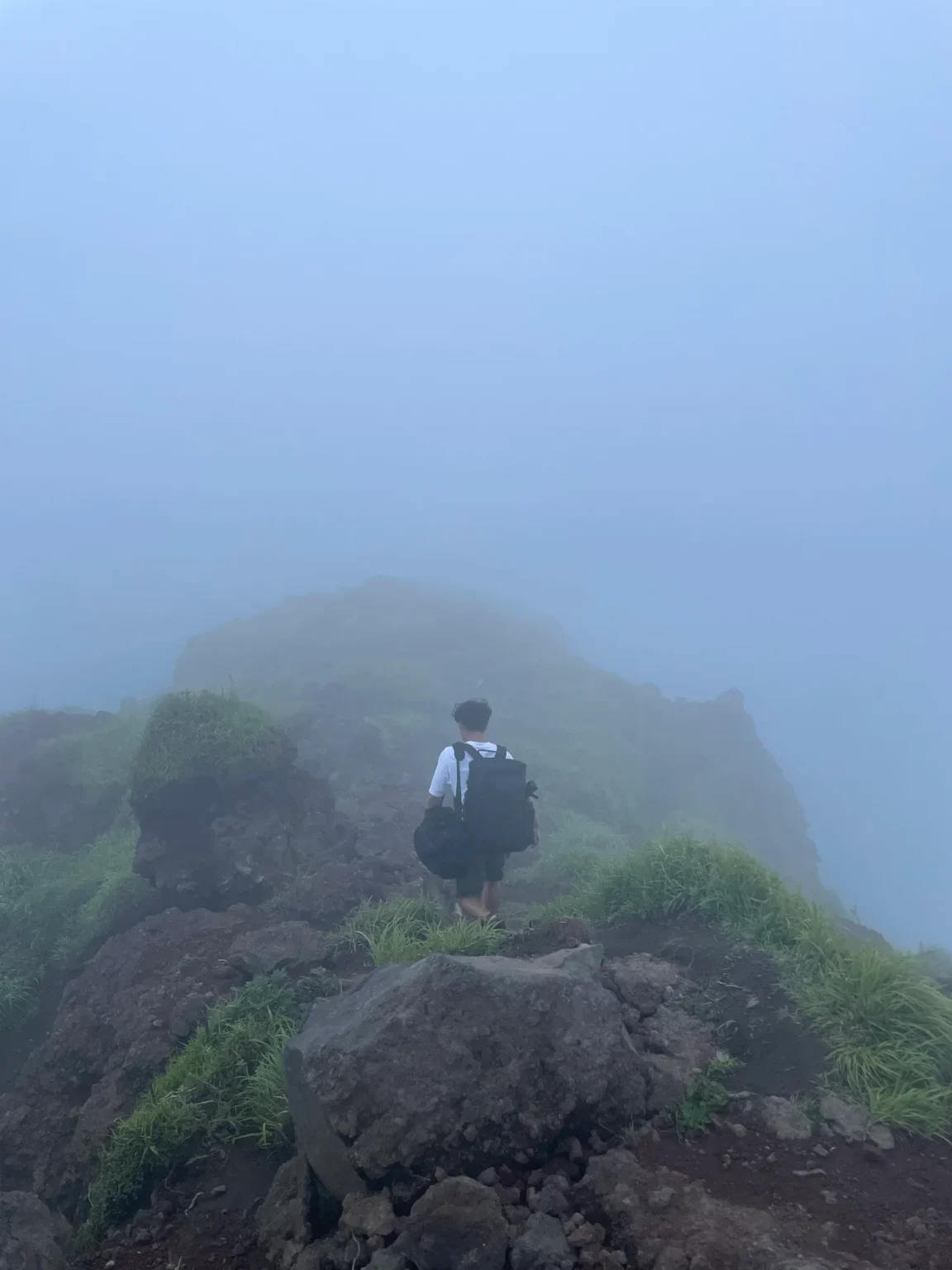 A person with a black backpack, seen from behind, walks along a rocky, grassy ridge into thick grey fog; the outline of a higher rocky knoll is faint ahead.