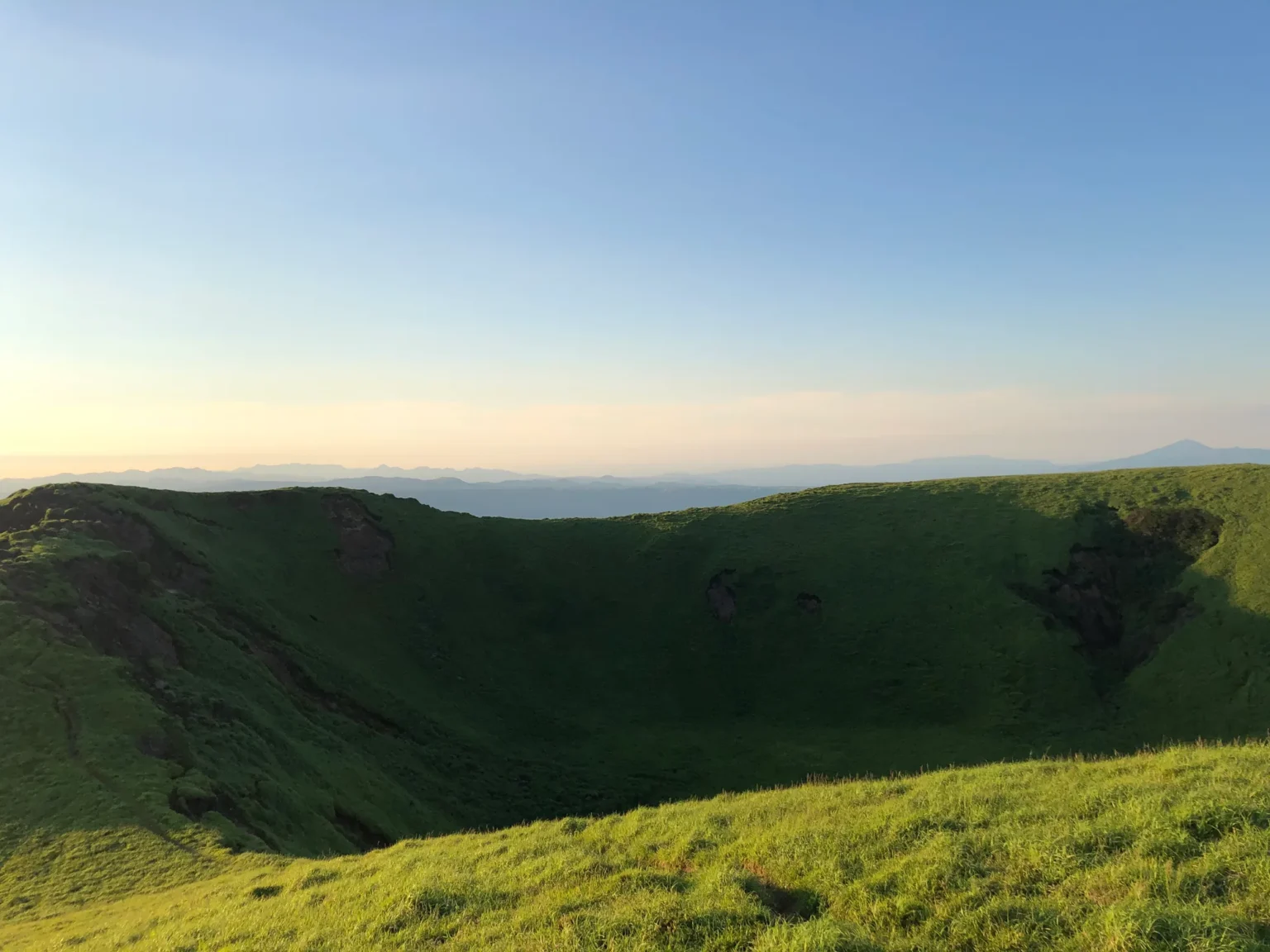 A deep bowl-shaped crater lined with green grass, partly in shadow, with patches of dark rock on its inner walls; beyond the far rim, hazy hills and a distant peak under a pale evening sky.