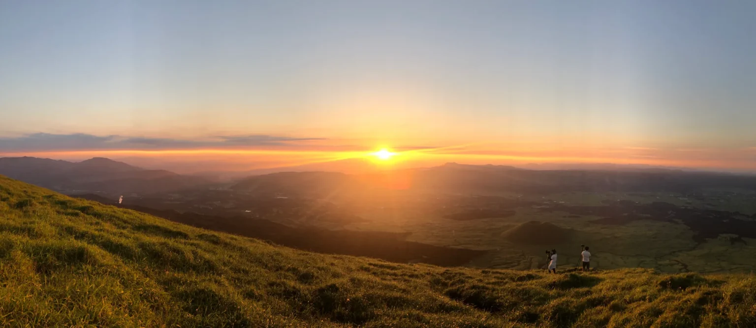 Wide panorama at sunset from a grassy slope: the sun low over the caldera's western rim, the green caldera floor below, a small round cone with a hollow top at lower right, and two people with cameras on tripods standing on the slope in the foreground.