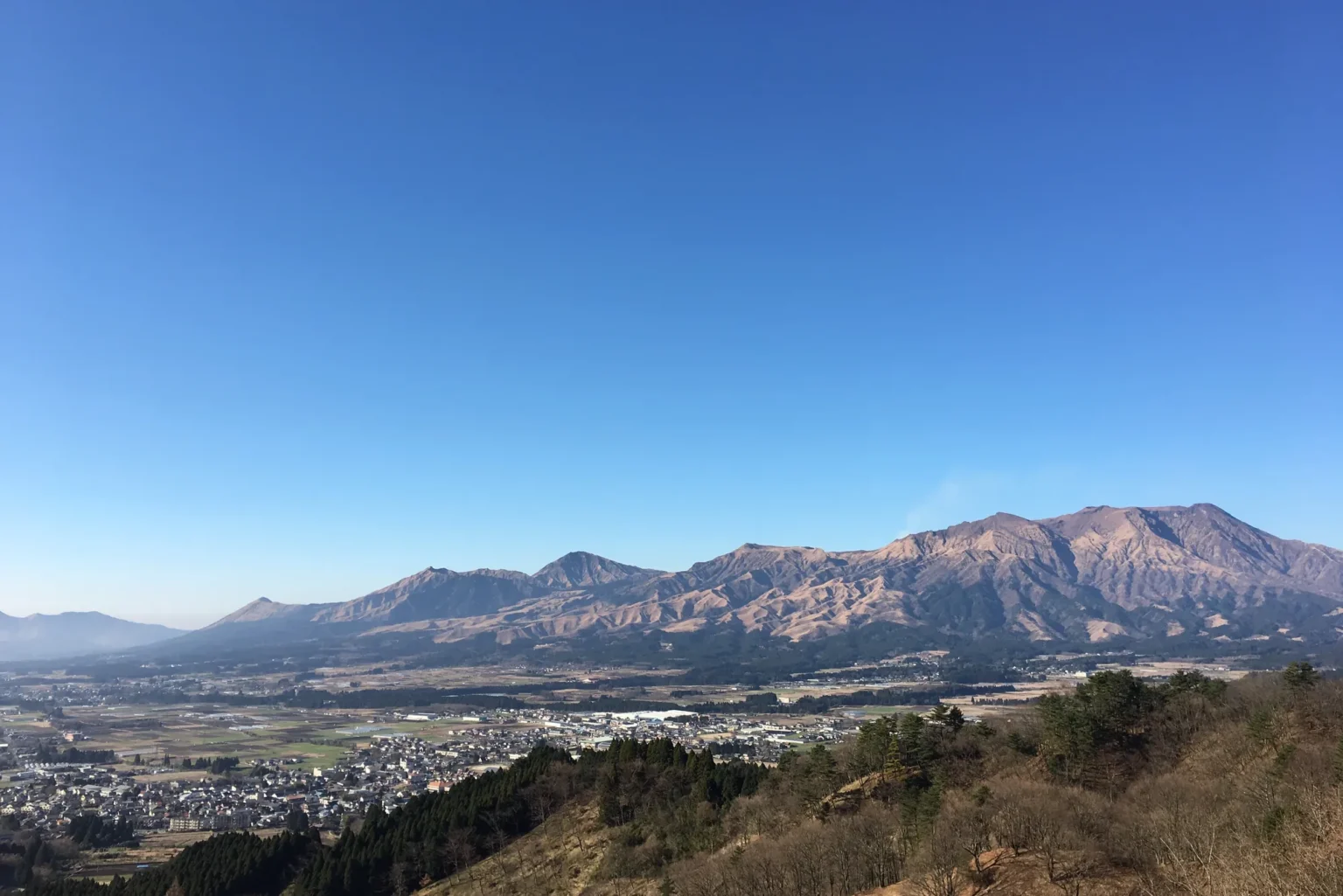 Under a clear blue sky, a long jagged mountain range with brown, deeply gullied slopes rises above a wide valley with a town of low buildings and fields; a wooded hillside in the foreground.