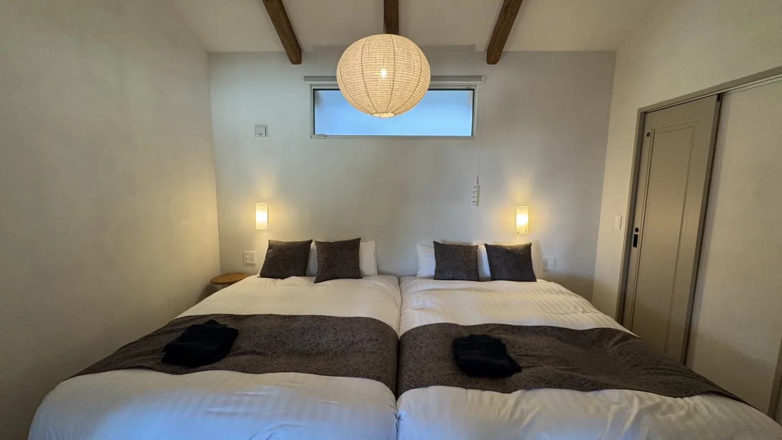 A bedroom with two beds pushed together, a paper pendant lamp, and a small high window