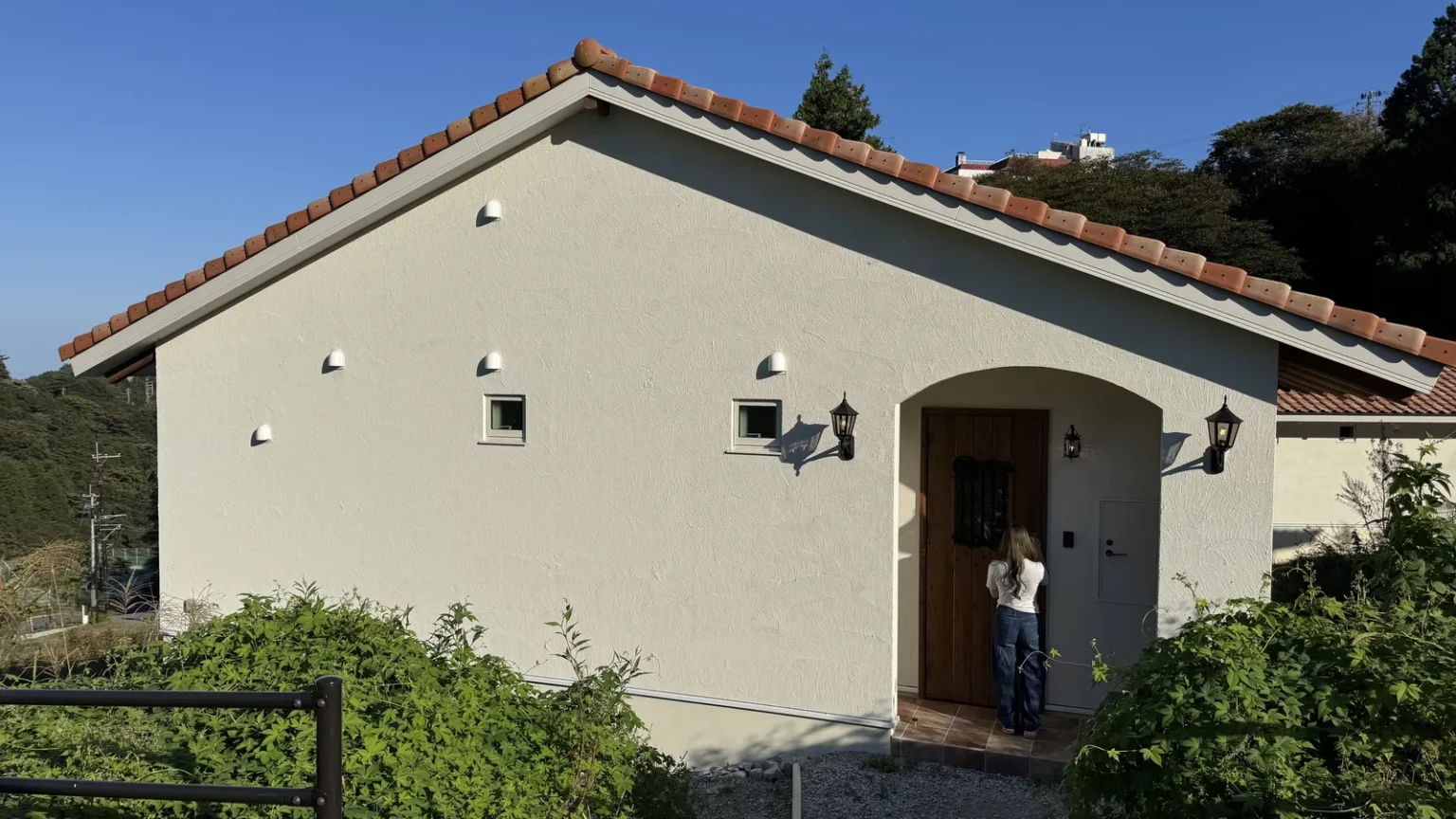 A white single-storey cottage with a terracotta tile roof and an arched wooden front door, a person at the door, green shrubs and a blue sky