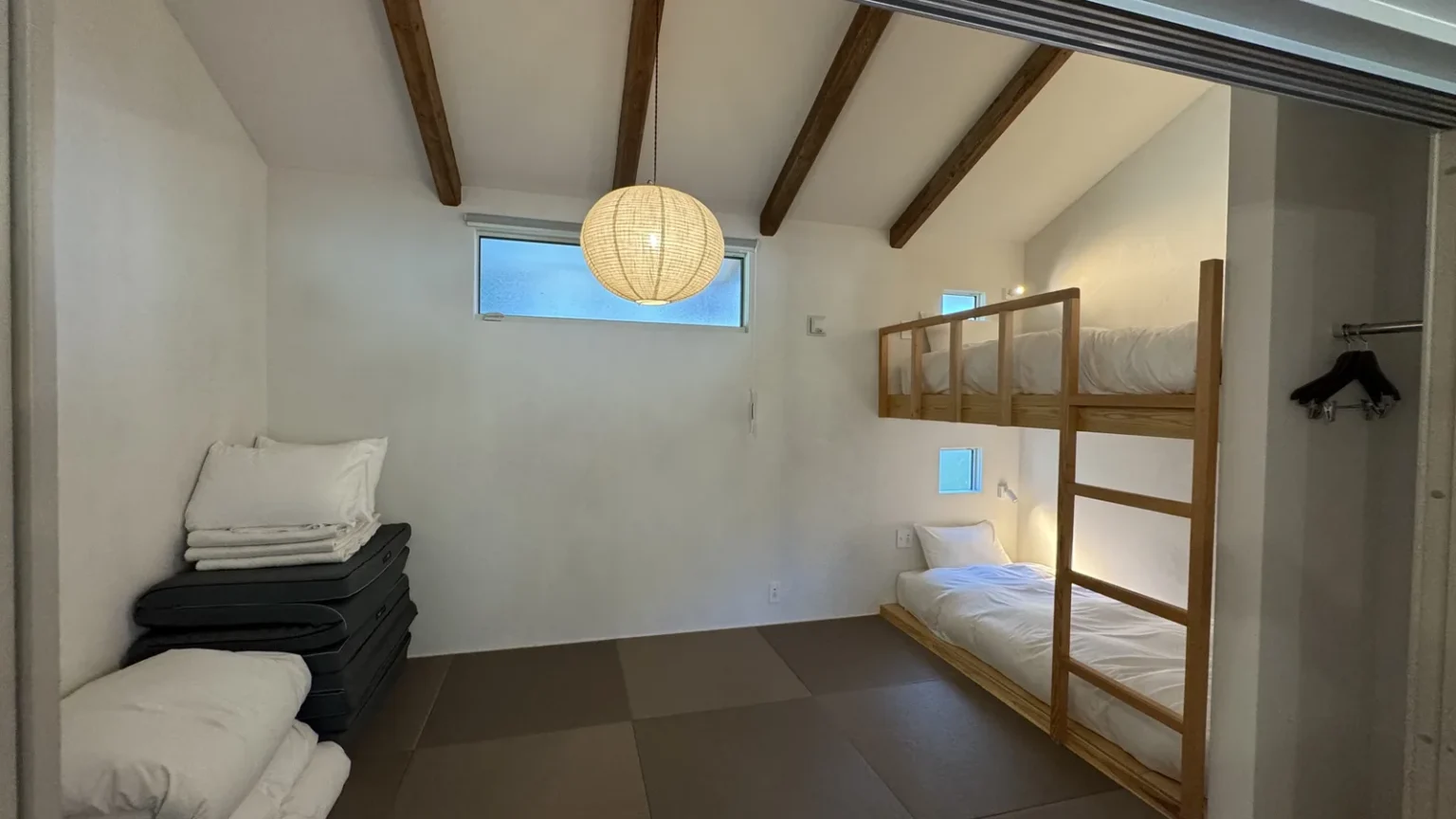 A room with tatami-style flooring, stacked futons and pillows, and a wooden bunk bed against the wall