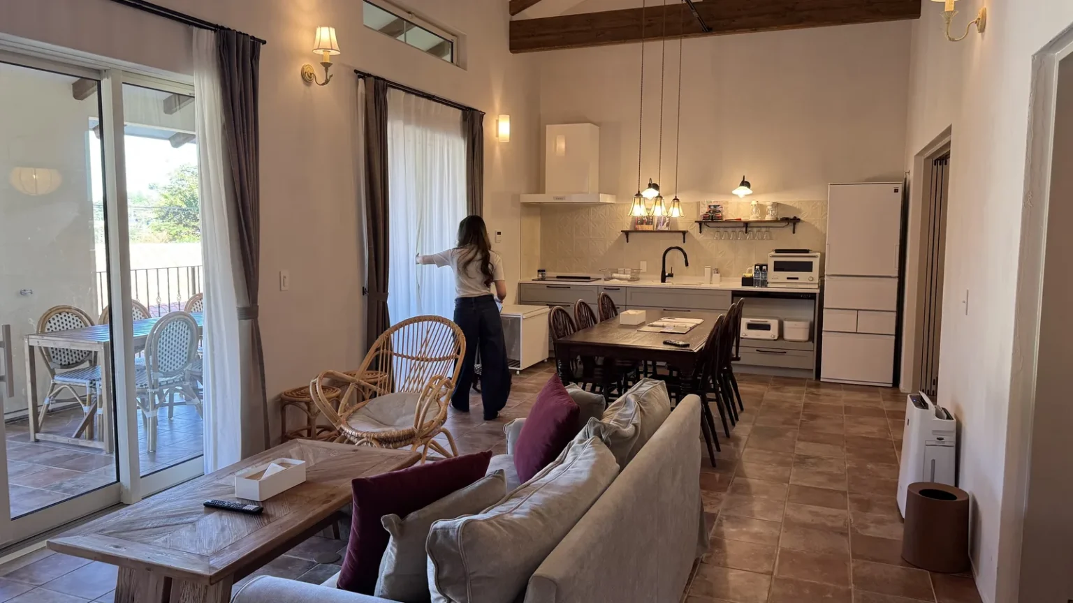 A living room with a grey sofa, rattan chair and wooden coffee table, a long dining table and an open kitchen with a large fridge, under a high beamed ceiling, with a person by the window