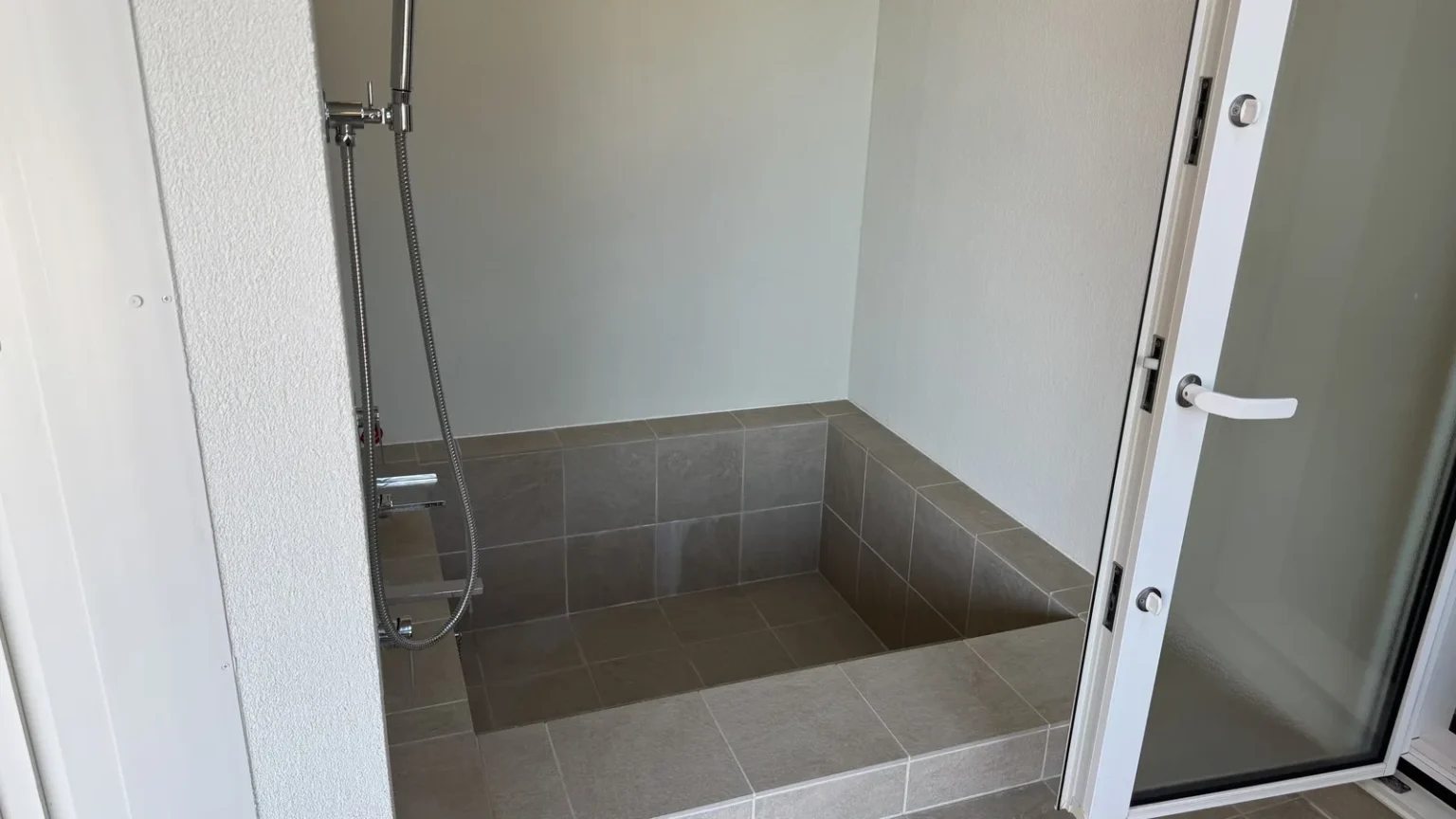A square tiled bath sunk into the floor beside a white wall, with a hand shower and an open glass door