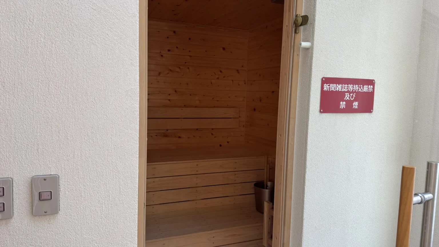 The open door of a small sauna with wood-panelled walls and wooden benches, next to a red sign in Japanese on a white wall