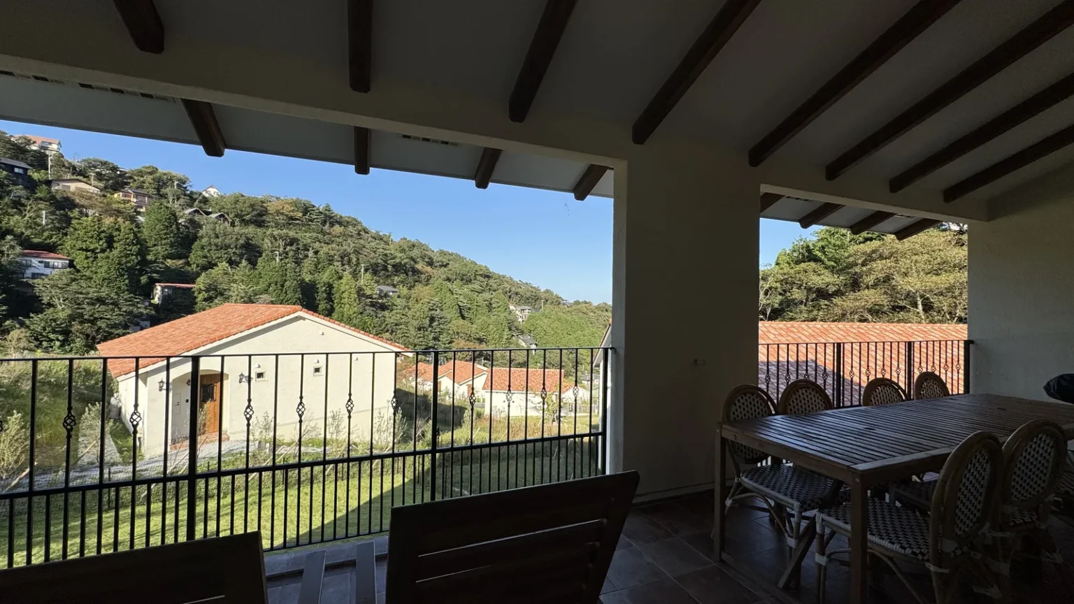 A covered terrace with a dining table and chairs behind a black railing, looking over a lawn to another white cottage with a red roof and green hills