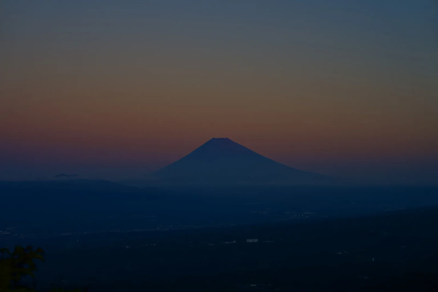 The dark silhouette of Mt Fuji against an orange and grey dawn sky, above a dark plain
