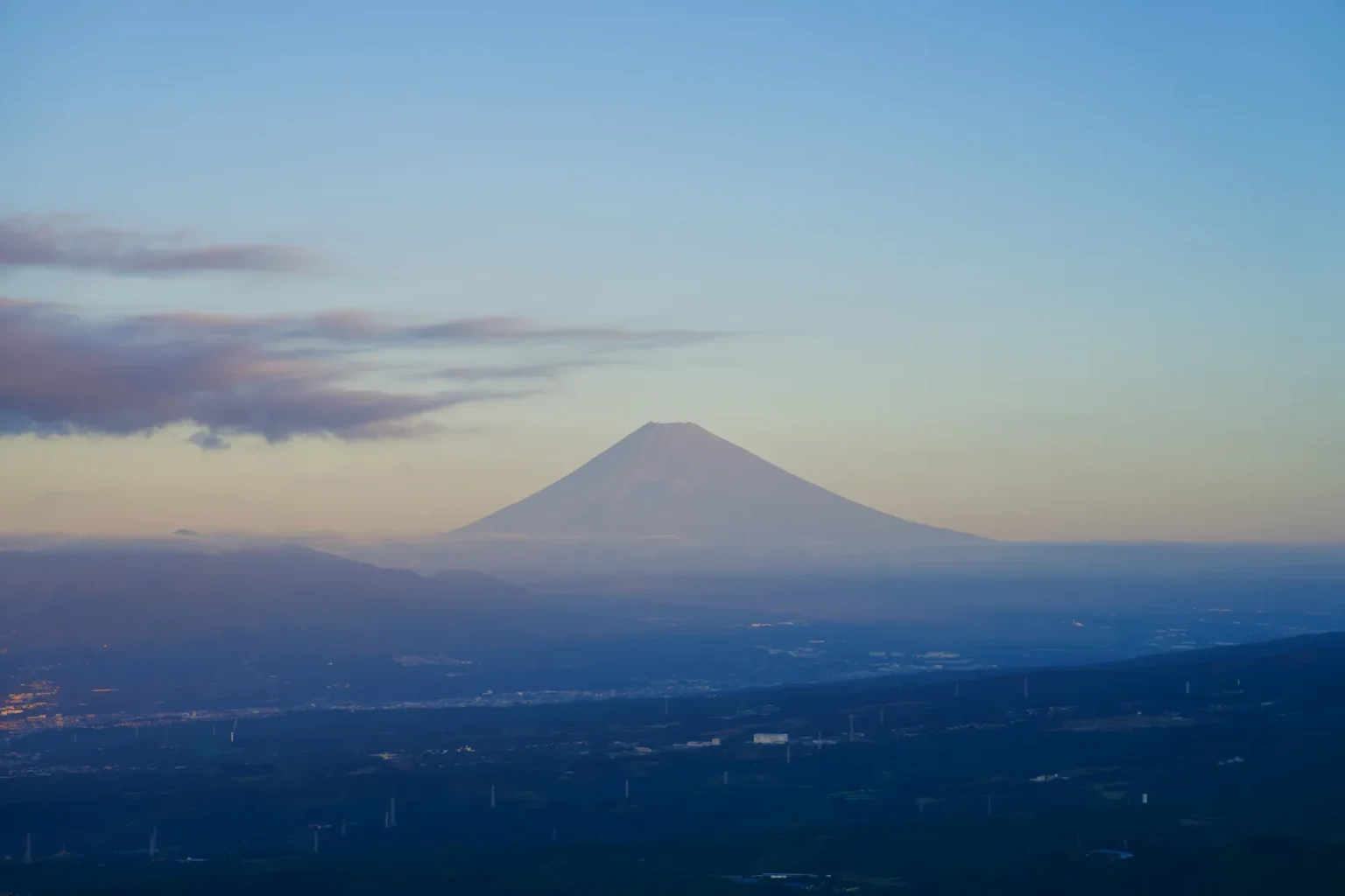 Mt Fuji under a pale blue sky with streaks of cloud, above a wide plain and low hills