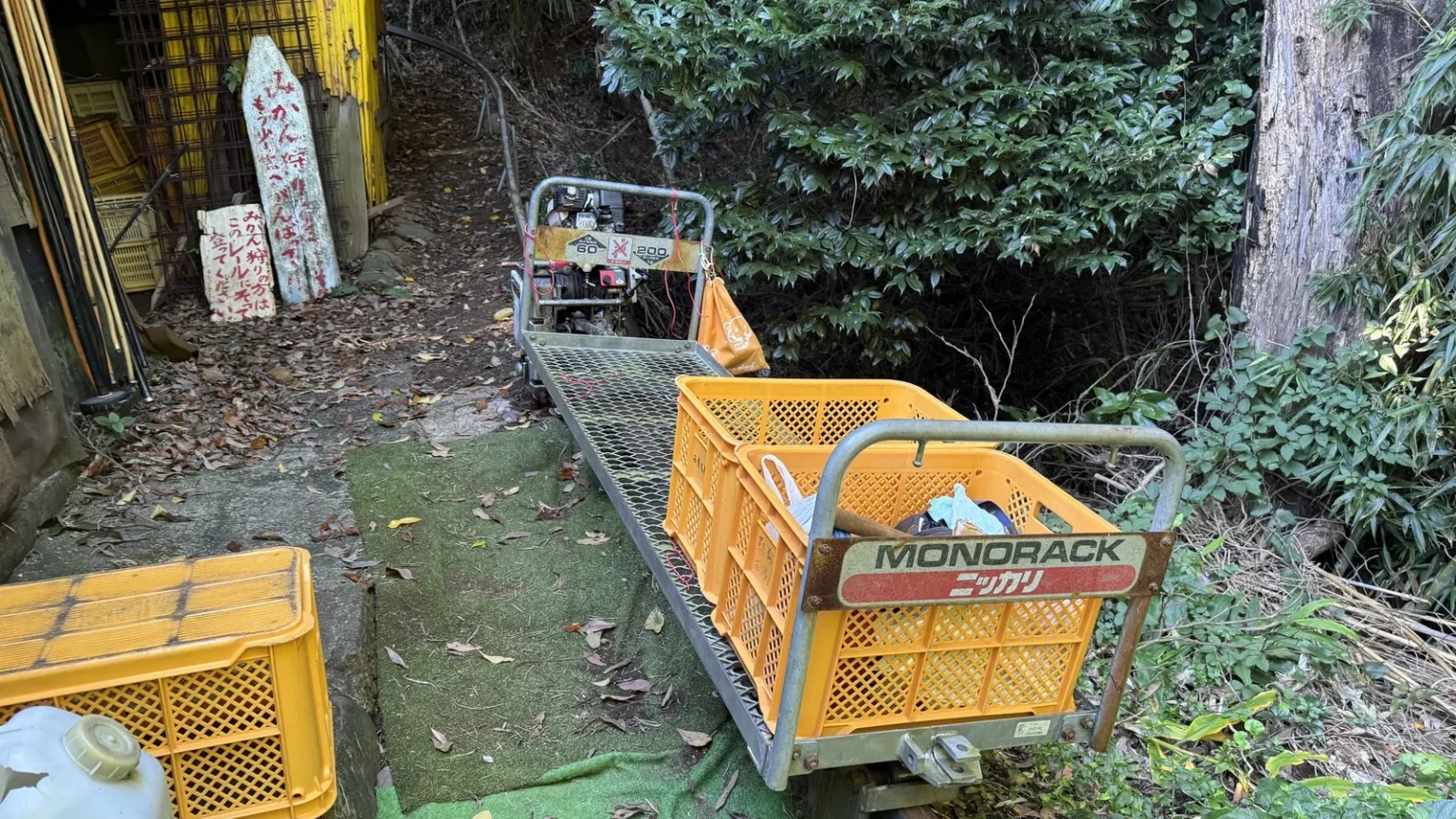 A small farm monorail cart labelled Monorack carrying an orange plastic crate on a rail, beside more crates and fallen leaves