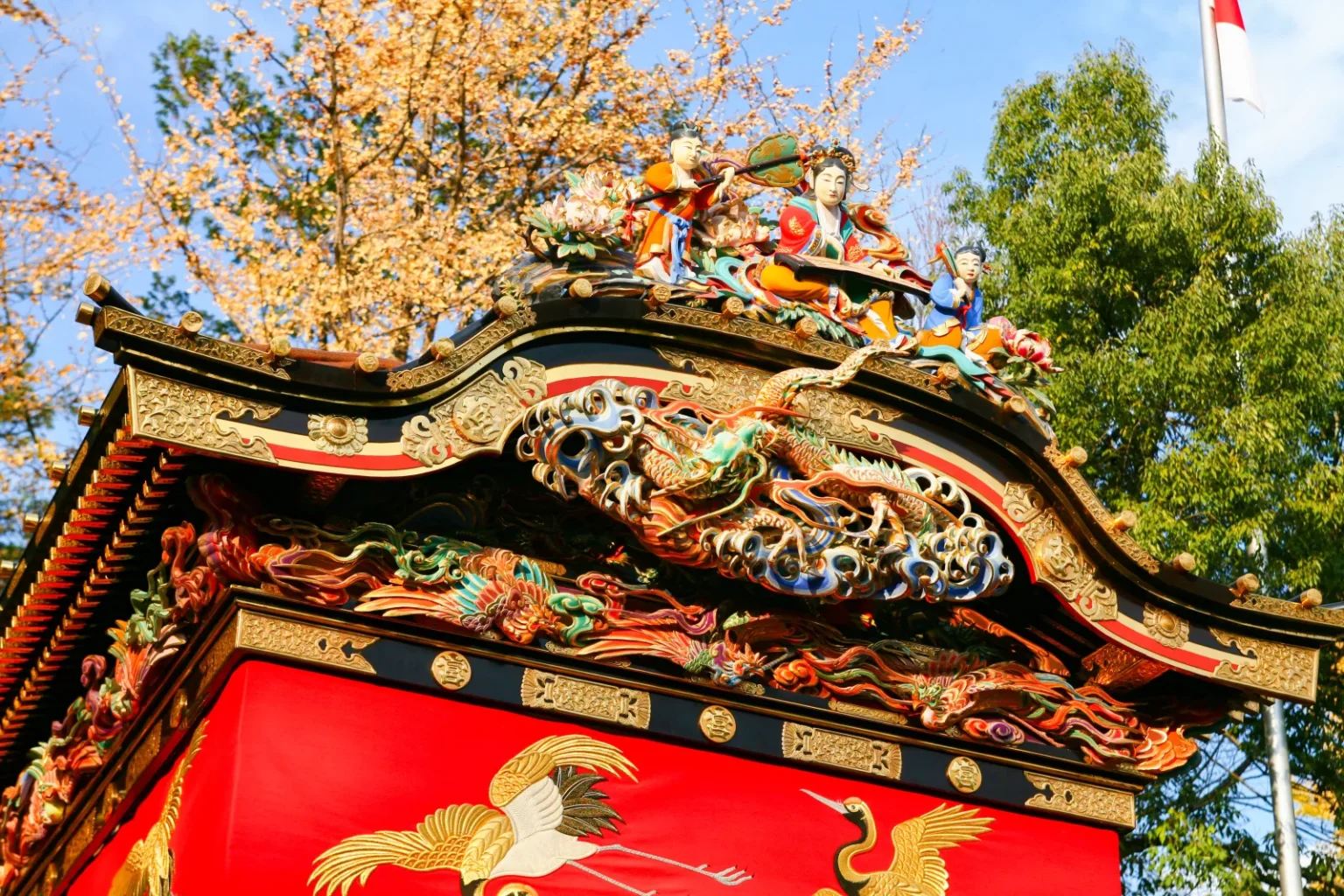 Close view of the ornate carved and painted roof of a festival float, with red panels showing cranes, against trees and sky