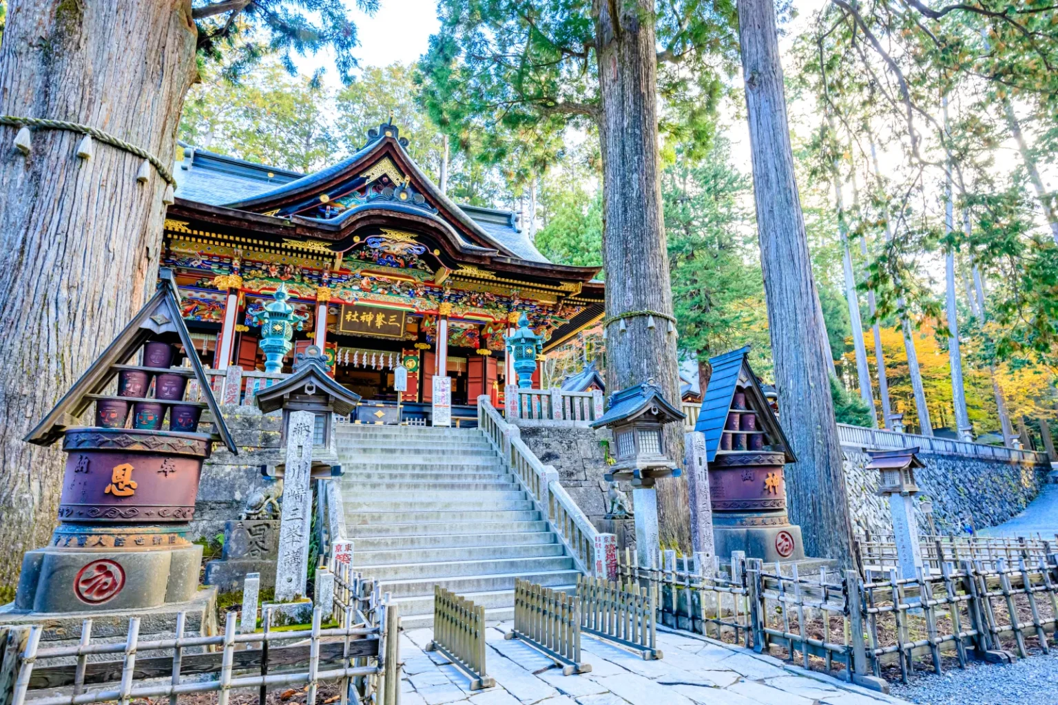 Stone steps rising to an ornate shrine hall among tall cedar trees, with lanterns and autumn leaves