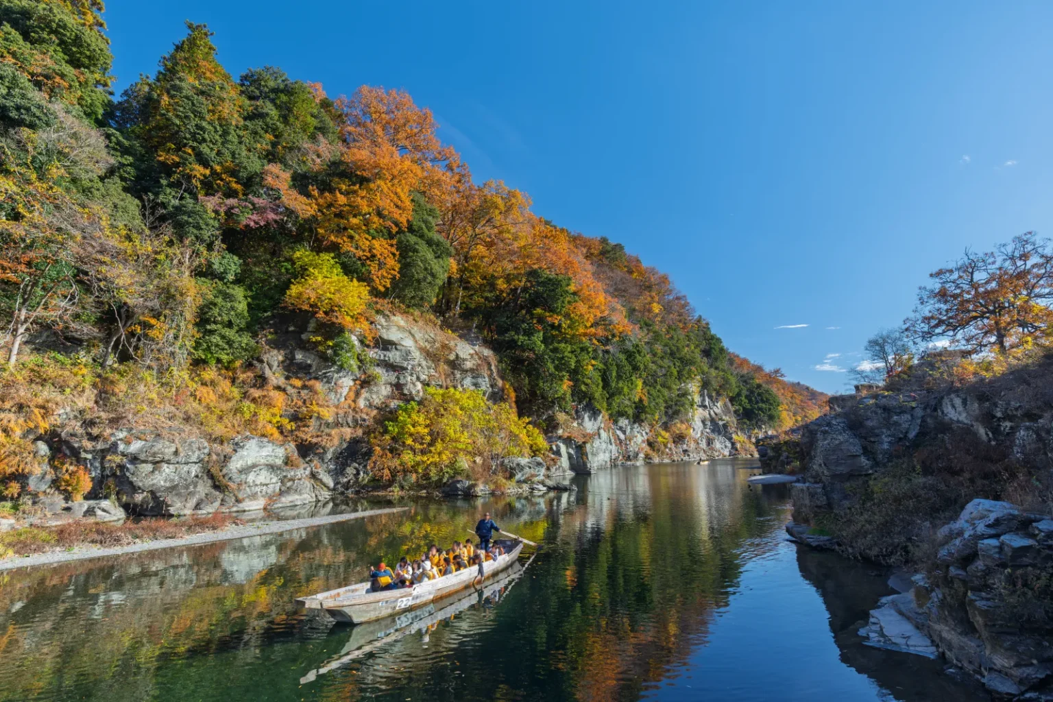 A long wooden boat with passengers on a calm green river between rocky banks and autumn-coloured hills under a blue sky