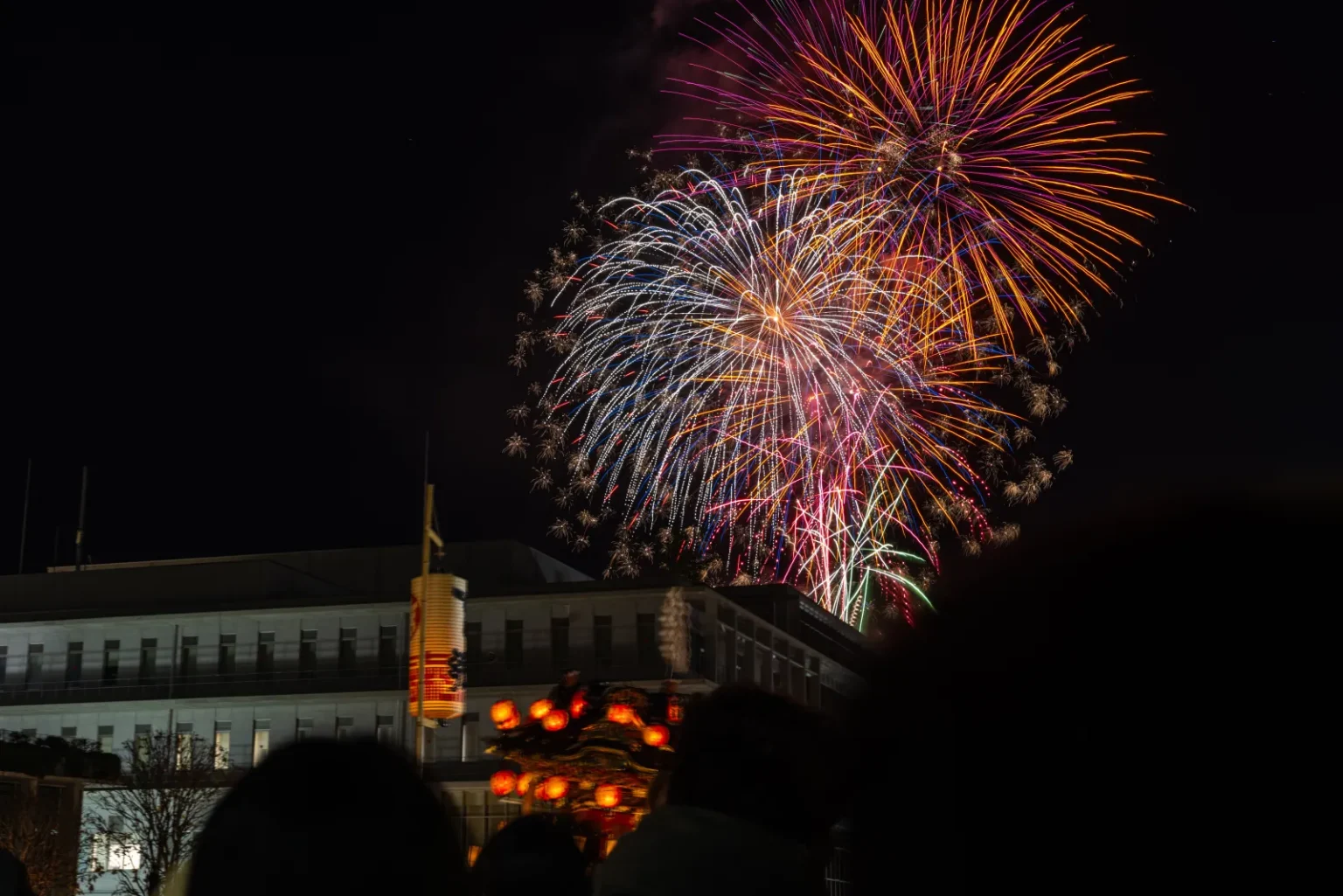 Fireworks bursting in the night sky above a lit festival float and a building, with silhouettes of people in the foreground
