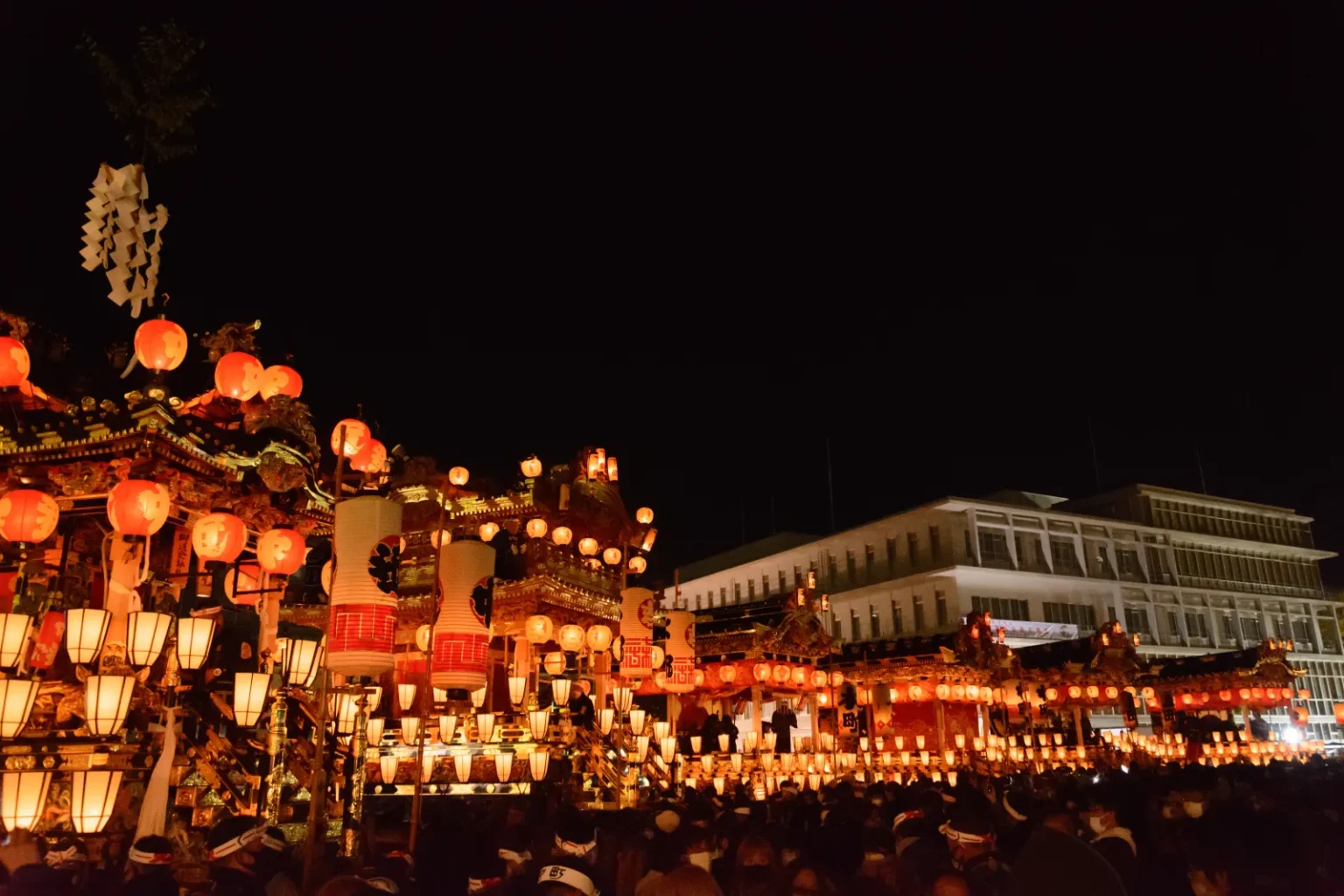 Festival floats lit with rows of red and white lanterns at night, with crowds and a building behind