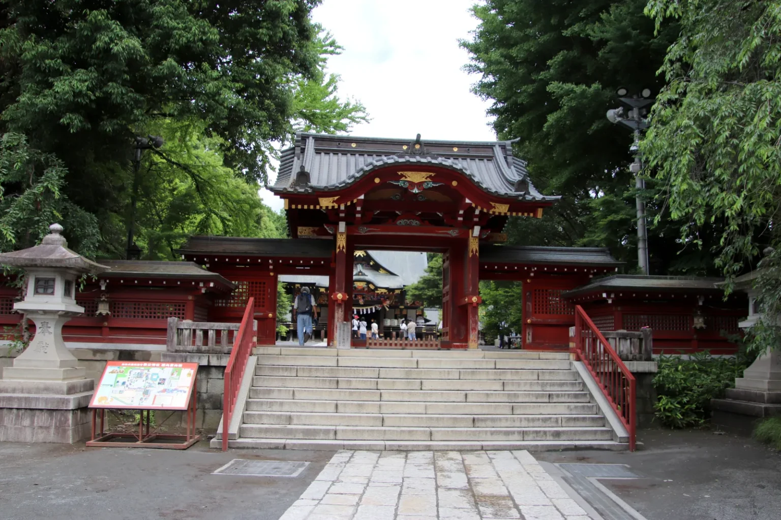 A red two-storey gate at the top of stone steps, with stone lanterns, a sign board and tall trees around it