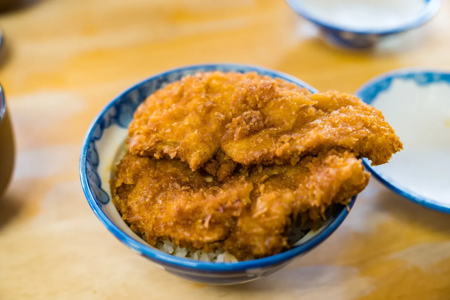 A blue-and-white bowl of rice topped with two large breaded pork cutlets, on a wooden table