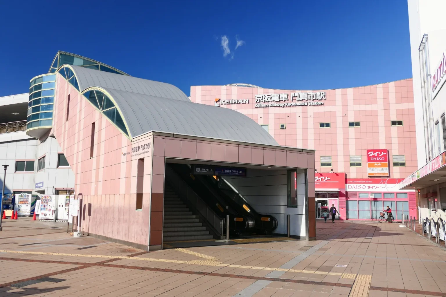 The pink-tiled south entrance of Keihan Kadomashi Station with escalators and stairs under a curved grey roof, the station name in Japanese and English on the building behind, under a blue sky