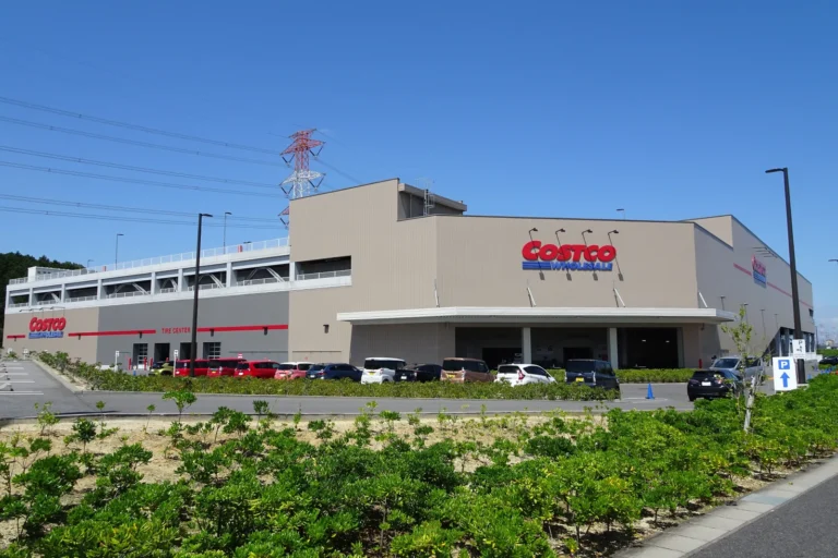 A large beige Costco Wholesale warehouse building with red Costco signs, cars parked in front and a multi-storey car park on its left, under a clear blue sky with shrubs in the foreground