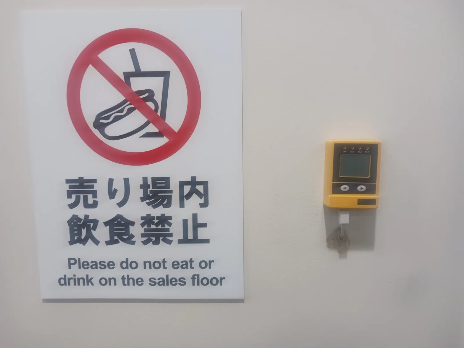 A white wall sign with a red no-entry symbol over a hot dog and a drink, reading in Japanese and English: Please do not eat or drink on the sales floor