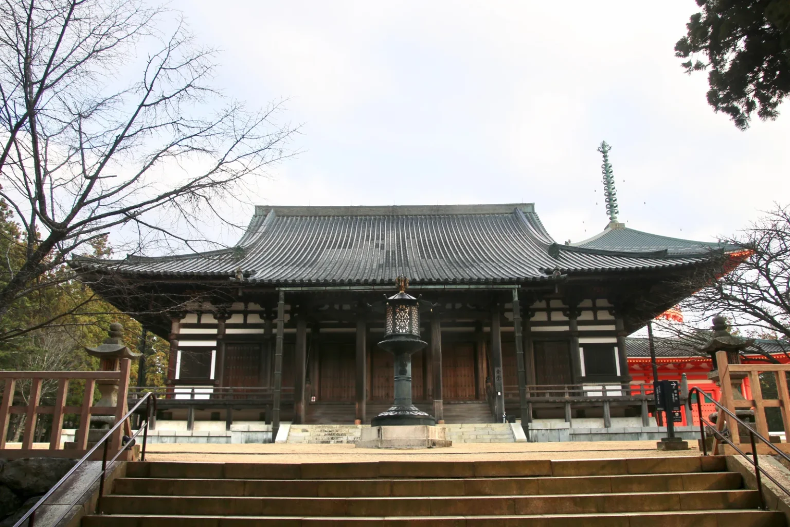 The Kondo main hall of Danjo Garan with a wide grey hipped roof and a bronze lantern at the top of stone steps, with the red wall and finial of the Daito at right and bare winter trees