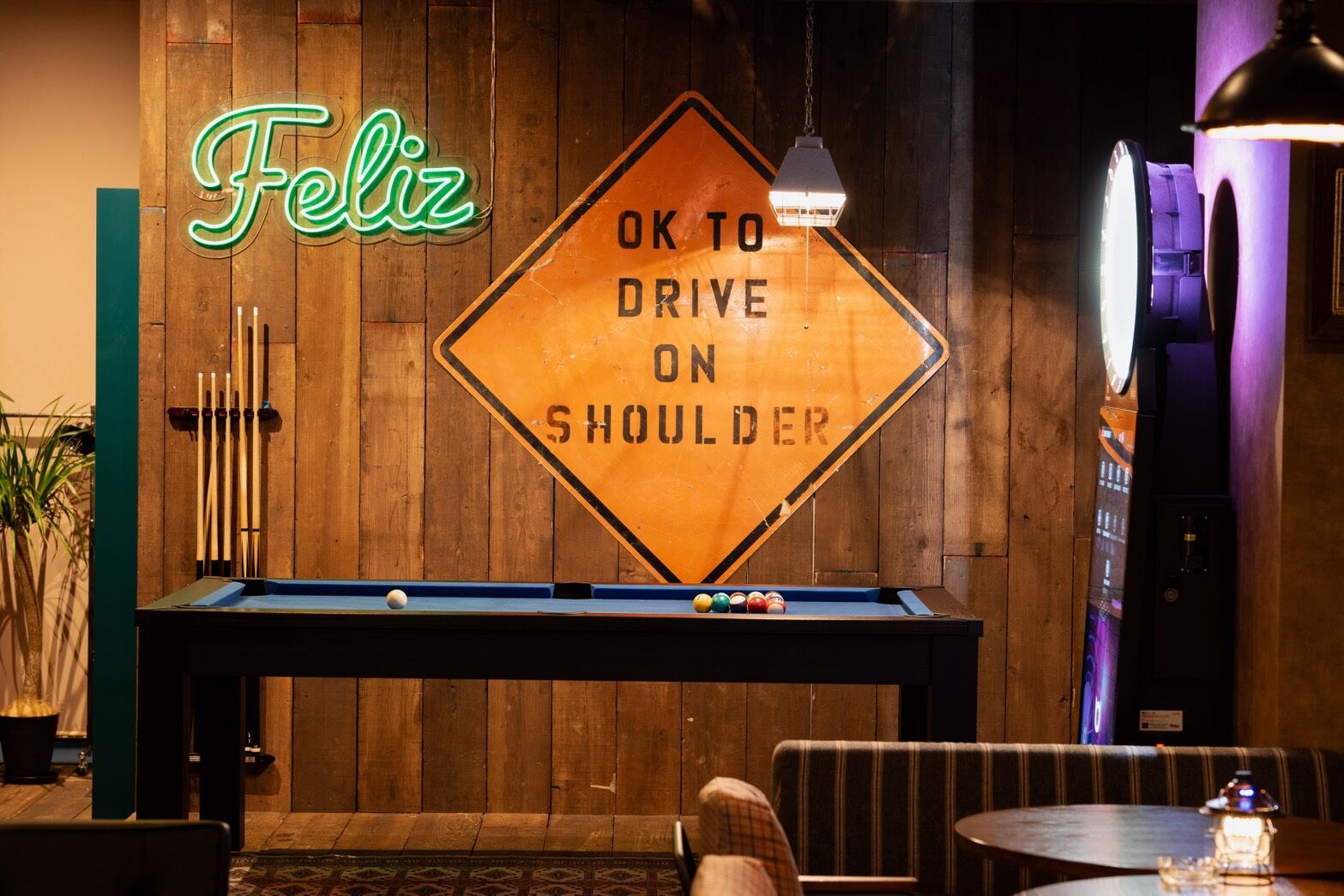 Wood-panelled room with a pool table, green neon Feliz sign and an orange road sign