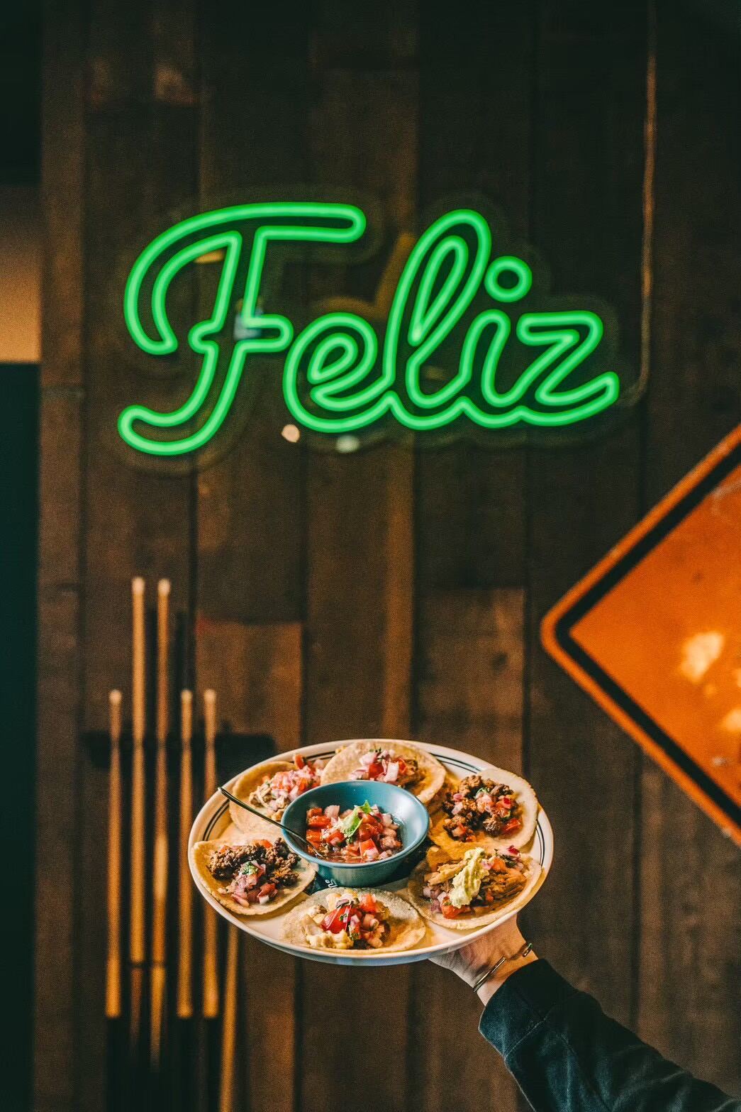 A green neon Feliz sign on a wooden wall above a plate of tacos