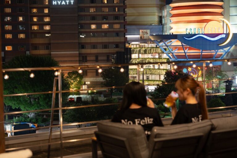 Two people on a rooftop terrace at night looking at lit-up hotel and building signs across the river