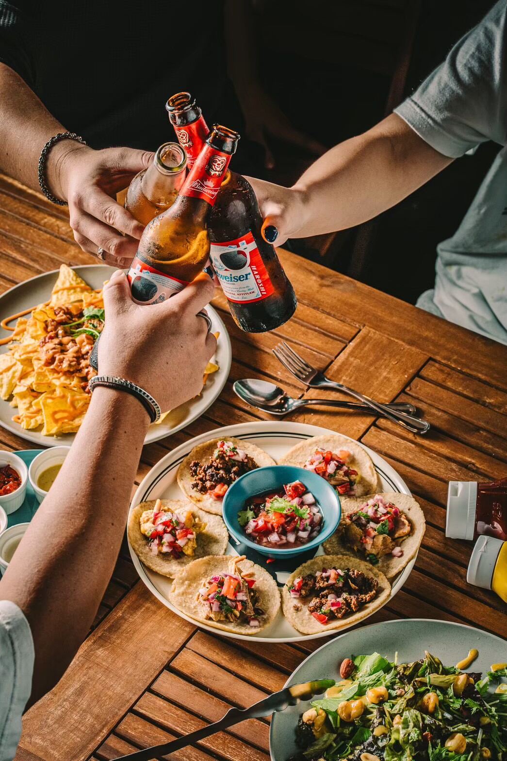 Hands clinking beer bottles over a table of tacos, salsa and salad