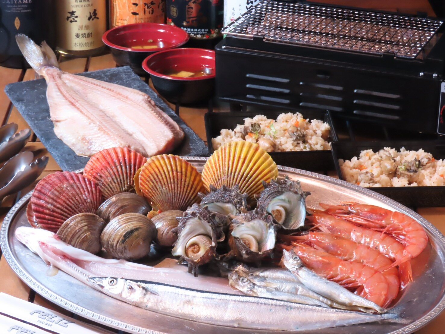 A platter of scallops, clams, turban shells, prawns and saury, with a split fish on a separate slate beside a tabletop grill