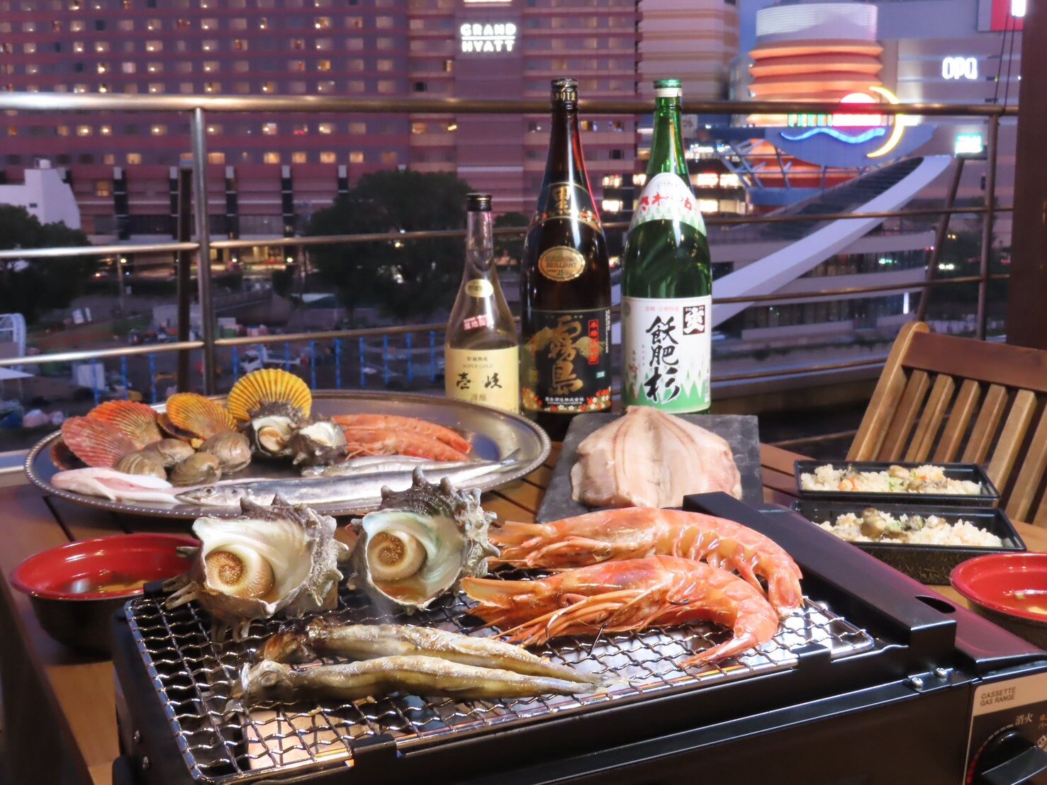 Turban shells, prawns and small fish on a tabletop grill on the rooftop at night, with lit hotel buildings behind