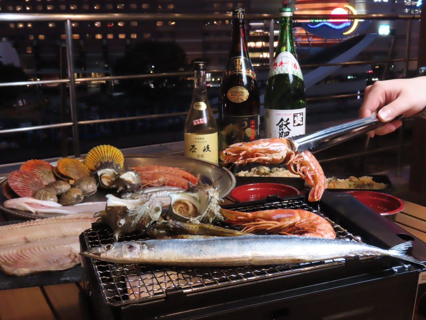 Tongs holding a prawn above a tabletop grill with saury and turban shells, with bottles of shochu behind