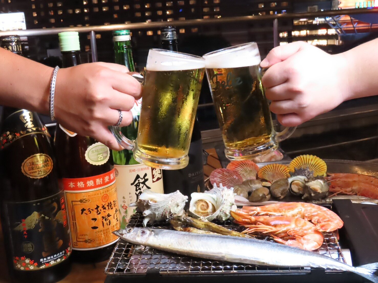 Two beer mugs clinking above a tabletop grill of saury, prawns and turban shells
